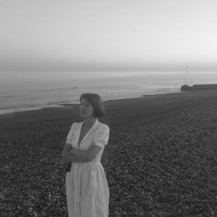 Betty standing on Brighton beach at dusk in a white tea dress with her arms crossed. 